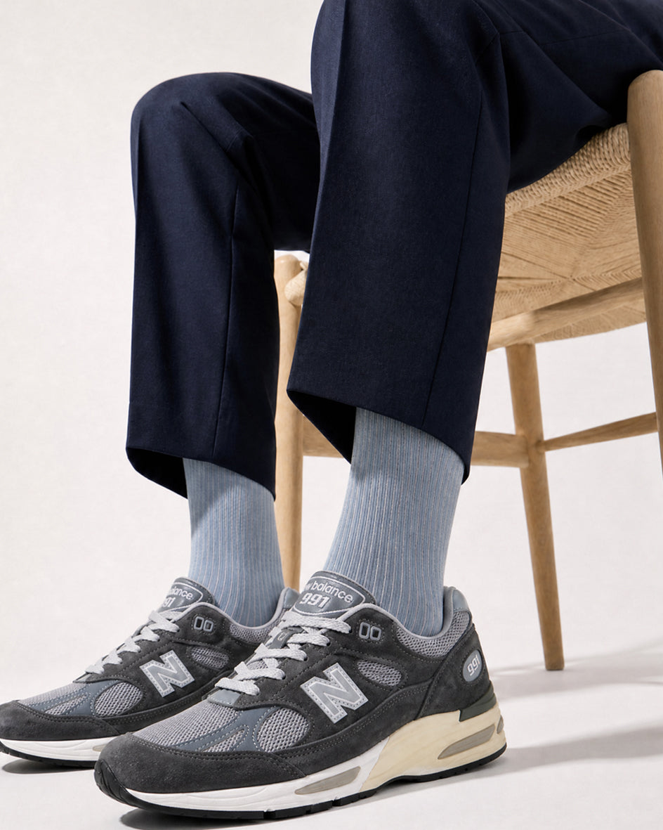 Light blue fine rib socks worn with navy trousers and grey New Balance 991 sneakers, seated on an oak paper-cord chair