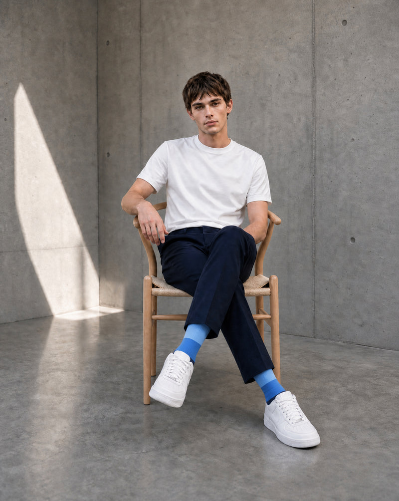 A man sits on a wooden chair in a minimalist concrete room, wearing a white crew-neck t-shirt, dark navy trousers, bright blue crew socks, and white low-top sneakers.