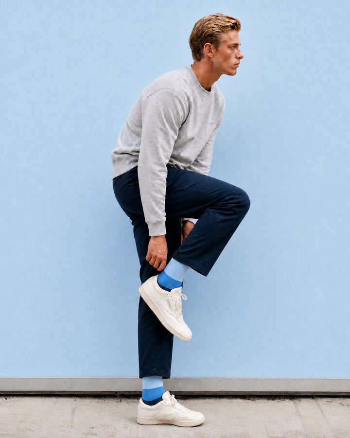 A man in a light gray crewneck sweatshirt, navy blue pants, white sneakers, and bright blue socks poses in profile against a solid light blue wall.